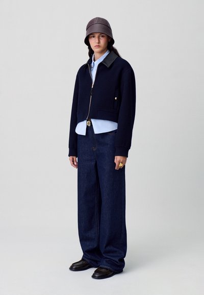 Chaqueta navy corta con cremallera, combinada con una camisa de color azul claro, pantalones de mezclilla oscuros anchos y zapatos negros. Acentuada con un sombrero de cubo marrón.