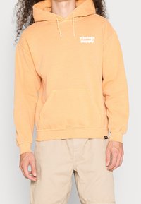 Orange hooded sweatshirt with a front pocket, "Vintage Supply" text in white, ribbed cuffs, and a relaxed fit. Fabric appears soft and slightly textured.