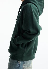 Personne portant un sweat à capuche vert foncé avec des poches avant et un jean en denim délavé clair, debout les mains dans les poches devant un fond blanc.