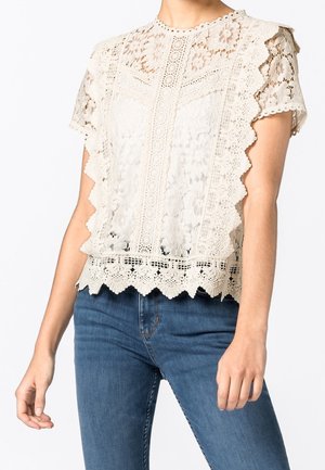 Vrouw in een crèmekleurige kantblouse met korte mouwen en bloemenpatronen, gecombineerd met blauwe denim jeans, staand tegen een witte achtergrond.