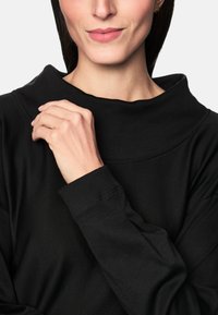 Frau trägt ein schwarzes Langarmoberteil, berührt mit der linken Hand den Kragen, Lippen geschlossen, und kleine goldene Creolen sind sichtbar.