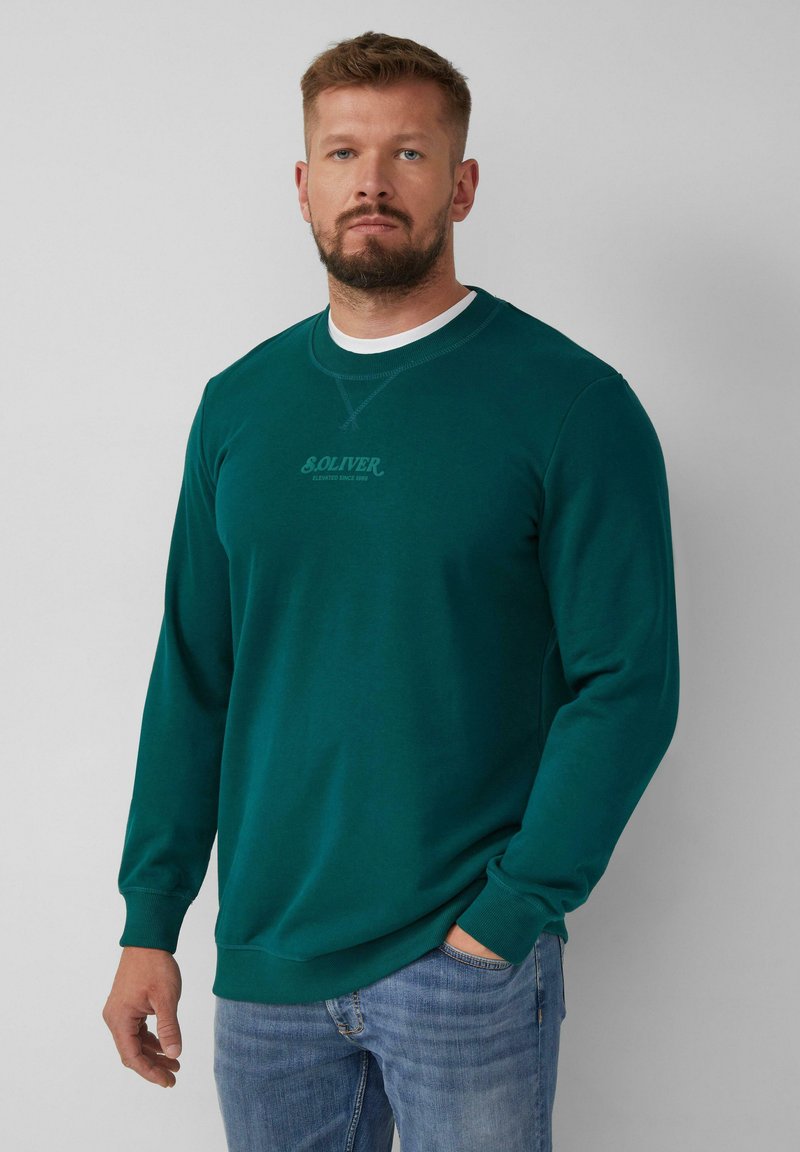 Felpa verde con maniche lunghe, scollo rotondo e dettaglio del logo sul petto. Indossata con jeans blu, che mostrano un look e uno stile casual.