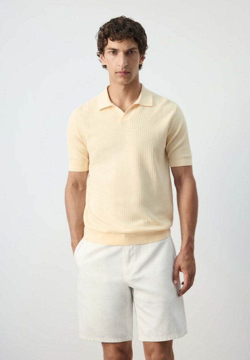Jeune homme aux cheveux bouclés portant un polo texturé à manches courtes jaune pâle et un short blanc, debout les mains dans les poches.