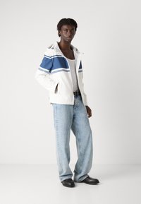 Hvid hættetrøje med blå striber, lynlås og lommer. Sammen med lyseblå wide-leg jeans og sorte sko.