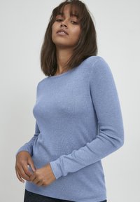 Svetlo modra dolgosleeve pulover s okroglim izrezom, rebrasto teksturo in prilegajočim oblikovanjem. Tkanina se zdi mehka in elastična.