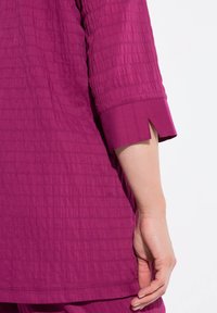 Magenta-toned teksturoidatud top, millel on kolmandiku pikkused varrukad ja jagatud disain ning sile äär. Kanga tekstuur on kortsus.