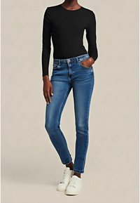 Personne portant un haut noir ajusté à manches longues, un jean bleu slim et des baskets blanches, debout avec une main dans la poche.