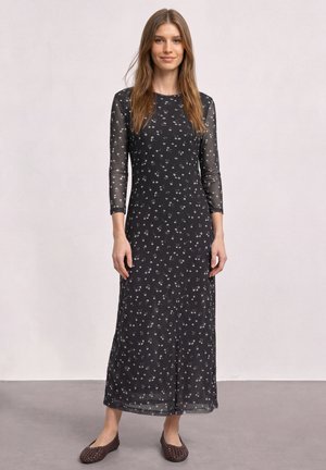 Frau steht und trägt ein schwarzes, langärmliges, transparentes Maxikleid mit floralem Muster, rundem Ausschnitt und braunen geflochtenen flachen Schuhen vor neutralem Hintergrund.