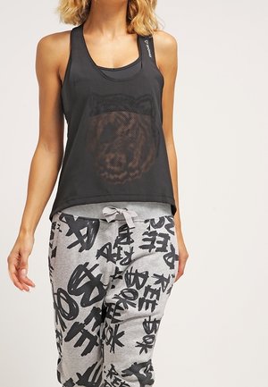 Vrouw draagt een zwart mesh tanktop met berengezicht print en grijze joggingbroek met zwarte graffiti-stijl tekst en een trekkoord in de taille.