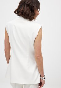 Bela brezrokavnik z strukturirano zasnovo, gladko teksturo in diskretno detajlno šivanko na hrbtu. Ima klasičen ovratnik in brez zapiral.