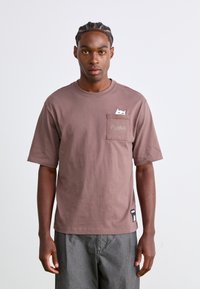 Tricou din bumbac de culoare maro, cu mâneci scurte, având o flamă cu un grafic de pisică albă și un logo "Puma" pe flamă.