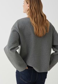 Grauer, übergroßer Pullover mit weiten Ärmeln und rundem Halsausschnitt, aus weichem Stoff. Kombiniert mit dunkelblauen Jeans, die einen leichten High-Low-Schnitt zeigen.