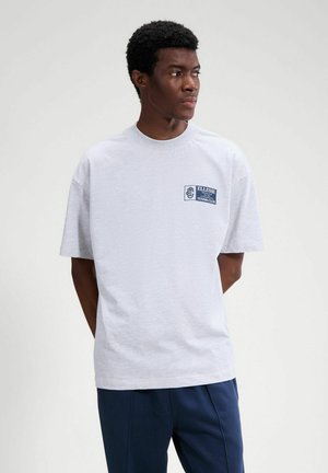 Giovane uomo con capelli corti e ricci, indossa una maglietta bianca con il logo blu "Ellesse Tennis Club" e pantaloni blu navy, guarda verso destra.