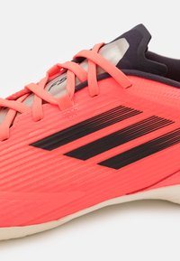 Bright coral soccer cleat with textured synthetic upper, black accent stripes, and flat laces. Features a low-cut, ergonomic design.