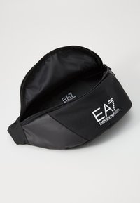 Sacoche noire avec fermeture éclair, arborant le logo EA7 bien en vue à l'avant. L'intérieur est partiellement visible.