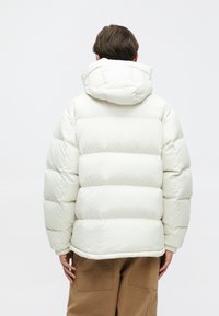 Doudoune blanche avec capuche, présentant des sections matelassées et des poignets élastiques. Portée sur un pantalon marron, mettant en avant un design minimaliste.