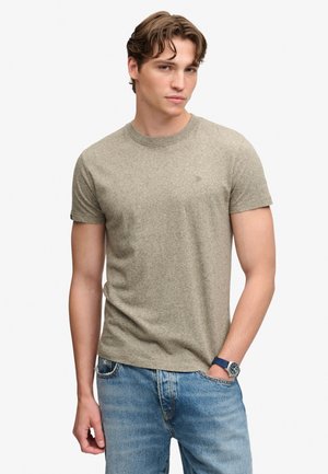 Junger Mann mit kurzen gewellten Haaren, der ein beiges T-Shirt mit kurzen Ärmeln, blaue Jeans und eine blaue Armbanduhr trägt, steht mit einer Hand in der Tasche.