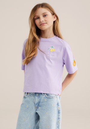 Joven niña con cabello rubio largo lleva una camiseta lavanda con bordado de frutas y unos vaqueros azul claro con pequeños estampados de flores.