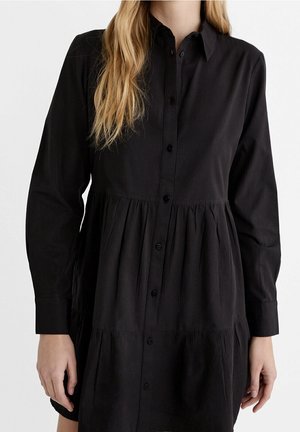 Vrouw draagt een zwarte jurk met lange mouwen, knopen, kraag en een geplooide taille, staand tegen een effen witte achtergrond.