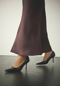 Mustat lakatun nahan korkokengät, joissa on terävä kärki ja hoikka piikkikorko, yhdistettynä liehuvaan tummanruskeaan mekkoon.