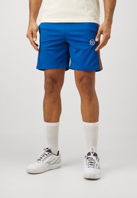 Sinised spordilühikesed, mis on valmistatud kerge kangaga, omavad tikandiga logo ja kontrastseid oranže külgriba. Kandsid koos valgete põlvini sokkidega.