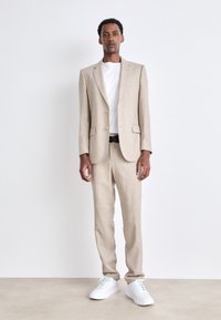Beige Leinenanzug mit einreihiger Jacke und passender Hose. Getragen über einem weißen T-Shirt, ergänzt durch weiße Sneaker.