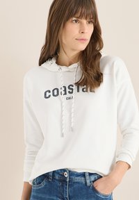 Sweat à capuche blanc avec un texte imprimé "coastal". Il est doté d'une capuche à cordon avec des accents argentés et est associé à un jean bleu.