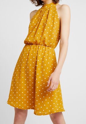Bezrekawna musztardowo-żółta sukienka w białe grochy, z halterowym dekoltem i marszczoną talią, noszona przez osobę z włosami do ramion.