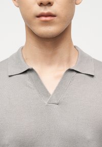 Gros plan sur le bas du visage et le cou d'une personne portant un pull en maille gris clair à col en V avec un col plat.