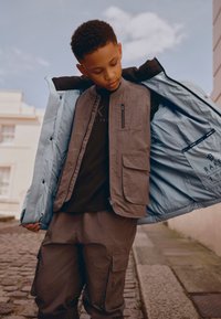 Niño al aire libre con pantalones cargo marrones y chaleco, sosteniendo abierta una chaqueta azul claro, mirando hacia abajo.
