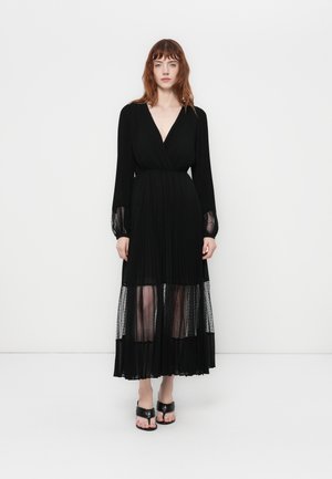 Woman wearing a long black pleated dress with sheer dotted panels at sleeves and hem, paired with black open-toe heels.