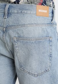 Los vaqueros de mezclilla azul claro presentan un bolsillo trasero, una etiqueta de cuero marrón con la marca "WEEKDAY" y detalles de costura visibles. Textura suave y corte casual.