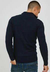 Marineblå striktr�øje med en ribbet krave, kontrastfarvede teksturerede kabelstrikkede ærmer og en tætsiddende kant, båret med jeans.