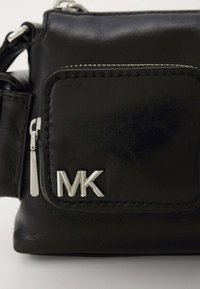 Bolso cruzado de cuero negro con una superficie texturizada. Cuenta con un bolsillo frontal con cremallera y un logo "MK" en tono plateado. Detalles de costura sutiles.