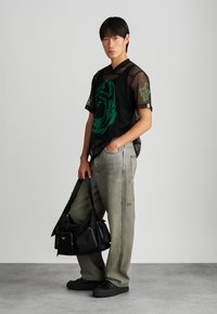 Camiseta de malha preta com design gráfico verde, combinada com jeans de perna larga cinza desbotados. Os acessórios incluem uma bolsa preta texturizada.