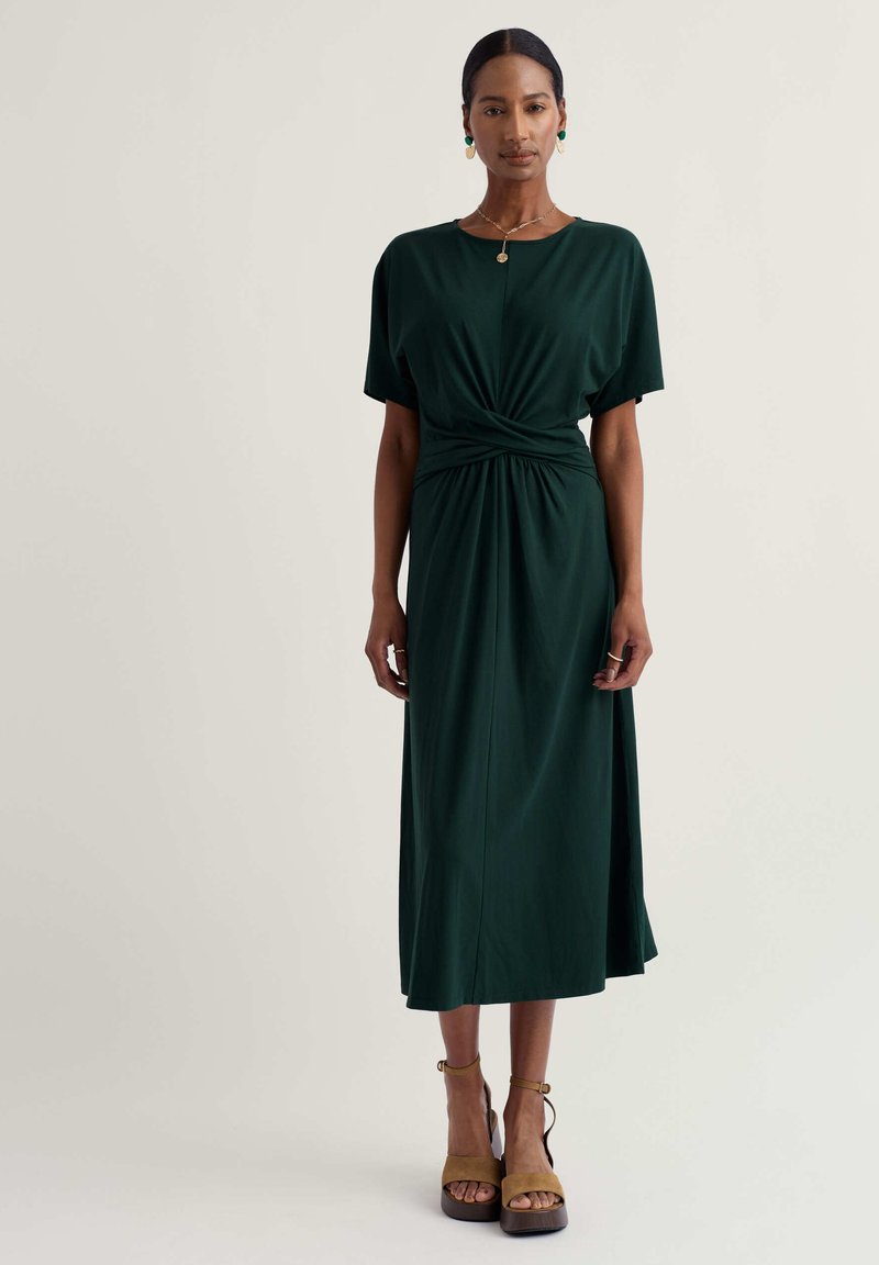 Femeie în picioare cu mâinile relaxate, purtând o rochie midi verde închis cu detaliu răsucit și sandale cu platformă maro deschis, pe un fundal deschis.