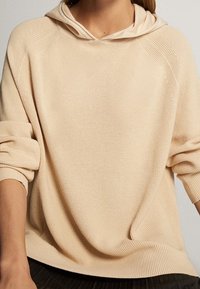 Sweat à capuche en maille beige avec une coupe décontractée, doté d'une grande capuche, d'une texture côtelée et d'épaules tombantes. Matière douce, design minimaliste.