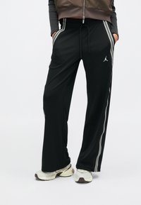 Persoon draagt zwarte wijde trainingsbroeken met witte zijstrepen, handen in de zakken, en witte sneakers, op een witte achtergrond.