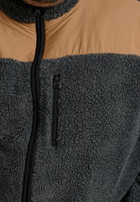 Hombre con chaqueta de forro polar negra y marrón con cremallera vertical negra y bolsillo con cremallera en el pecho visible.