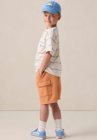 Un tout-petit portant une chemise rayée à manches courtes, un short cargo orange, des chaussures à carreaux bleus et une casquette bleue avec un design graphique.