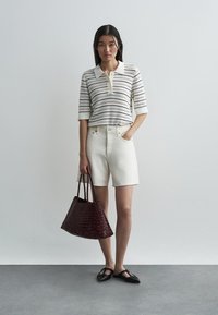 Adīts balts polo krekls ar melnām svītrām, baltas džinsi šorti, bordo krāsas austā soma un melni apavi uz pelēka grīdas.