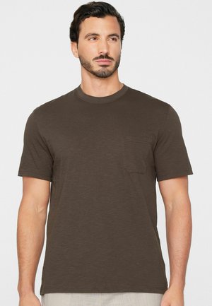 Mann mit kurzen dunklen Haaren und Bart, trägt ein einfarbiges dunkelbraunes kurzärmeliges T-Shirt mit Brusttasche vor weißem Hintergrund.