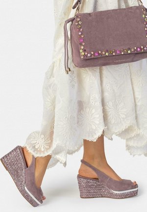 Vrouw draagt een witte geborduurde jurk met mauve suède sleehakken en bijpassende gestudeerde handtas, lopend op een witte achtergrond.