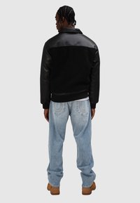 Czarna kurtka z teksturowanym panelem i skórzanymi rękawami, w połączeniu z jasnoniebieskimi, luźnymi spodniami dżinsowymi i brązowymi butami. Widok od tyłu.