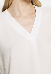 White, lightweight blouse with a v-neck design, featuring a smooth texture and short sleeves, highlighting a minimalist style.