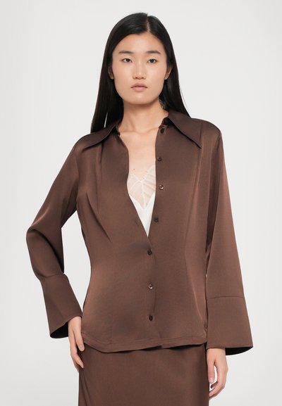 Brown silk button-up shirt with a wide collar and long sleeves. Features subtle sheen and structured fit. Worn over a light-colored top.