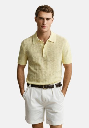Man draagt een lichtgeel gebreid poloshirt met korte mouwen en witte korte broek met een donker gevlochten riem, staat met zijn handen in de zakken.