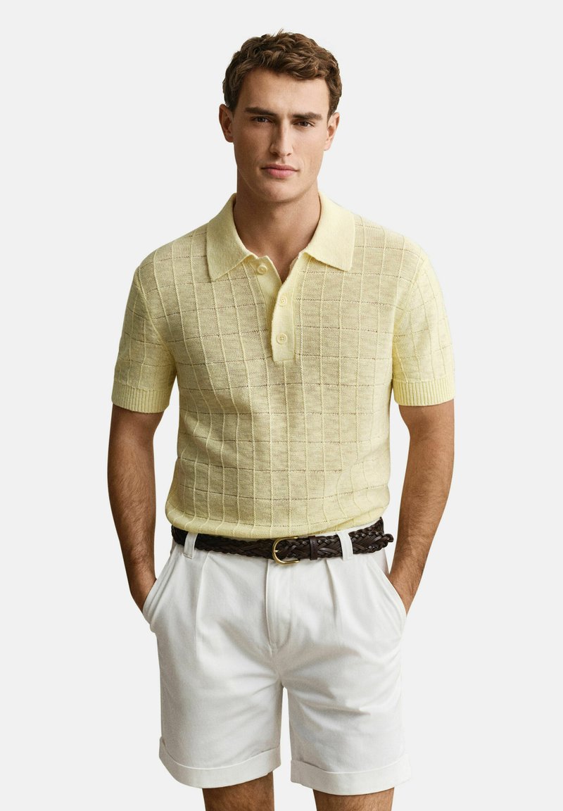 Mann trägt ein hellgelbes, kurzärmliges Strick-Poloshirt und weiße Shorts mit einem dunklen geflochtenen Gürtel, steht mit den Händen in den Taschen.