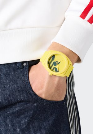 Dzeltena pulksteņa ar apaļu seju, kurai ir zaļa dizaina un stundu marķieru. Plastmasas josla un matēts apdare. Valkāta uz rokas, ar džinsu biksēm.