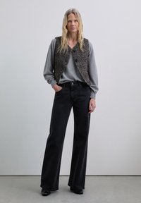 Camisa de manga larga gris, chaleco sin mangas con estampado, jeans de pierna ancha negros y zapatos negros. Texturas suaves y colores neutros.